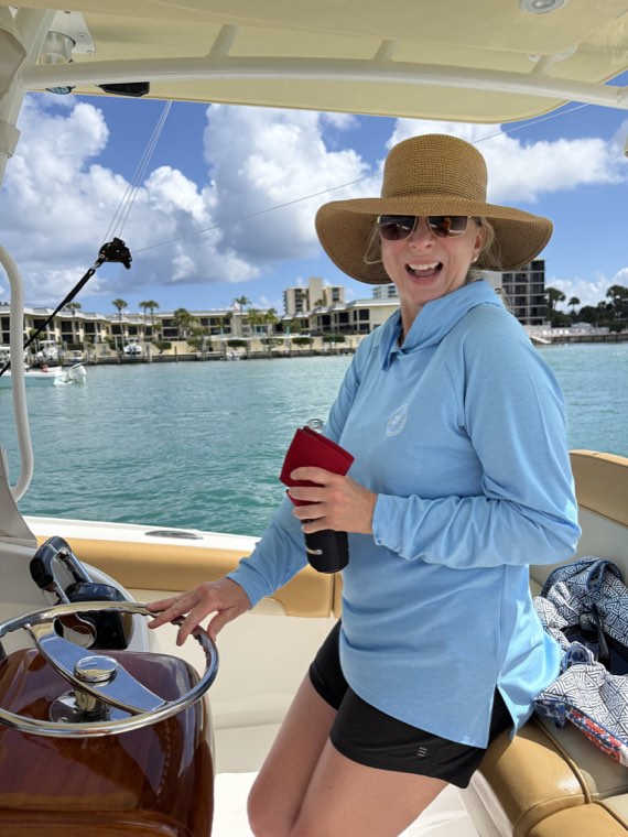 Courtney Nicholson at the helm of a boat in a sunlit marina, wearing a wide-brimmed hat.
