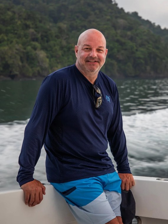 Jim Lucas standing at the rail of a boat on calm water, a forested shoreline behind him.