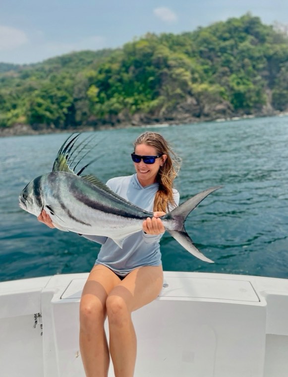 Rachael Shelton seated on a gunwale holding a roosterfish, a green coastline behind her.