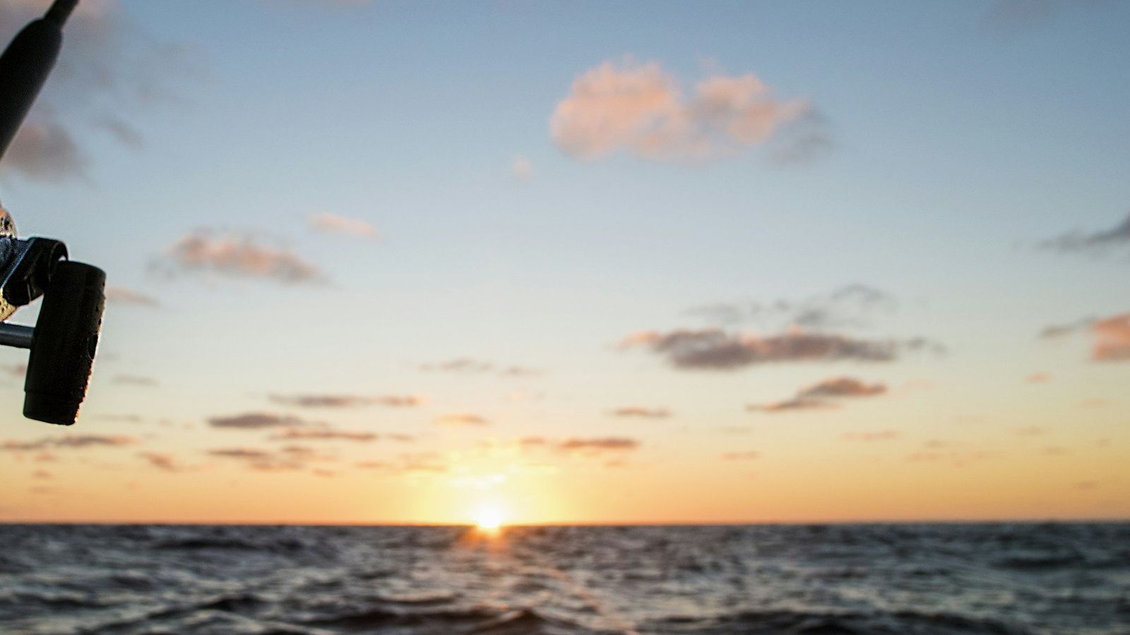 A trolling rod held steady in its holder as the sun sits on a level horizon over open ocean.