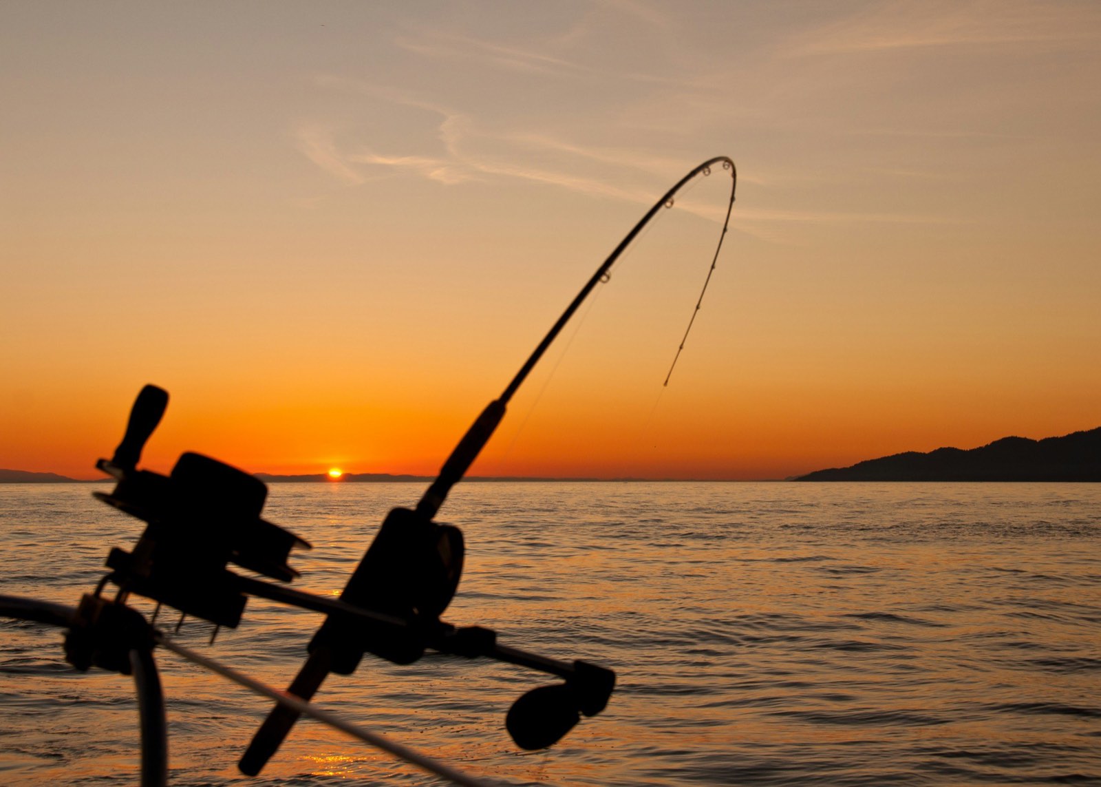 A bent trolling rod silhouetted against an orange sunset over open water.