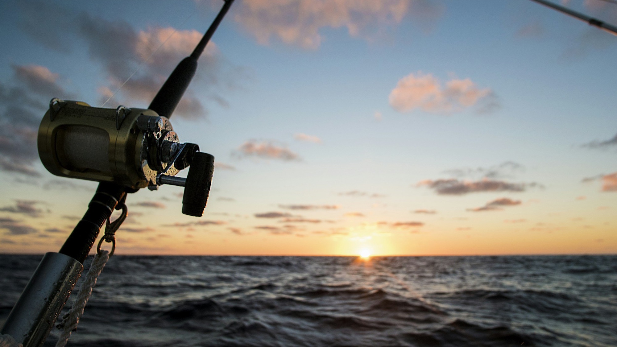A trolling reel mounted on the rail of a sportfishing boat as the sun sets over open ocean.