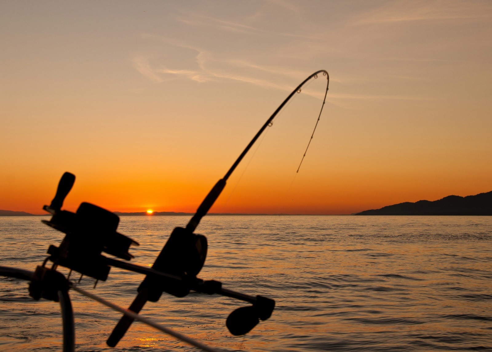 Silhouette of a fishing rod and reel set up on a boat during sunset over a body of water, with distant land on the horizon.